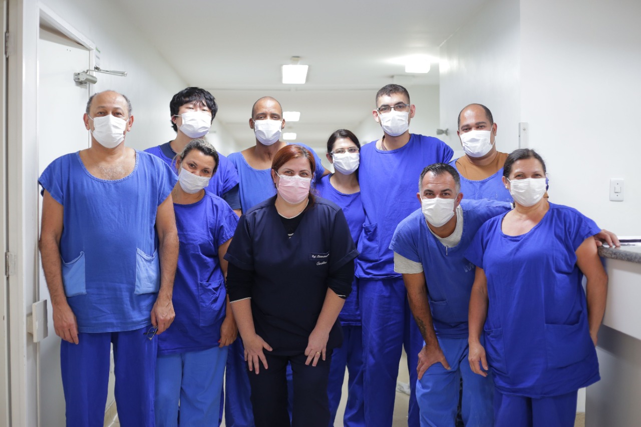 Na foto, Caroline está no meio de mais nove profissionais da saúde, dentre eles mulheres e homens. Todos estão em pé usando uniforme azul e máscara.